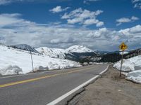 snow covers the roadway and snowy mountains on a sunny day, with a yellow warning sign in front of it