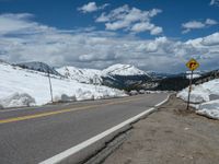 snow covers the roadway and snowy mountains on a sunny day, with a yellow warning sign in front of it