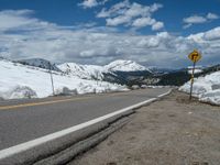 snow covers the roadway and snowy mountains on a sunny day, with a yellow warning sign in front of it