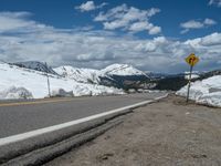 snow covers the roadway and snowy mountains on a sunny day, with a yellow warning sign in front of it