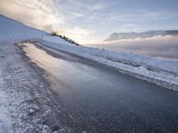 snow covered road beside large mountain in distance and sky in the background with clouds overhead