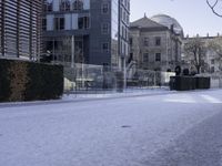 the sidewalk has snow in front of tall buildings and bushes covered in snow in front of a fence