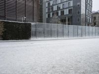 a snow covered street next to a tall building on the corner of a street with a fence