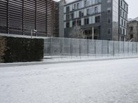 a snow covered street next to a tall building on the corner of a street with a fence