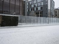 a snow covered street next to a tall building on the corner of a street with a fence