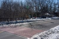 a street with a fence, cars and snow on it near a city sidewalk lined with trees