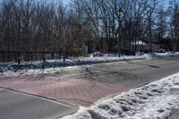 a street with a fence, cars and snow on it near a city sidewalk lined with trees