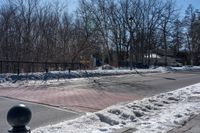 a street with a fence, cars and snow on it near a city sidewalk lined with trees