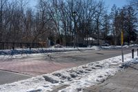 a street with a fence, cars and snow on it near a city sidewalk lined with trees