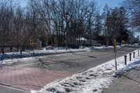 a street with a fence, cars and snow on it near a city sidewalk lined with trees