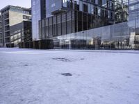 an empty area on a snow covered street surrounded by tall buildings and windows on one side