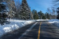 a winter street is paved with snow next to trees and a fence with pines