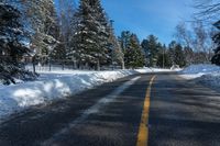 a winter street is paved with snow next to trees and a fence with pines