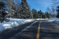 a winter street is paved with snow next to trees and a fence with pines