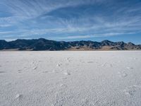 a snow field with mountains in the distance with clouds in the sky above it with footprints across the ice