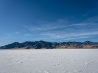 a snow field with mountains in the distance with clouds in the sky above it with footprints across the ice