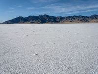 a snow field with mountains in the distance with clouds in the sky above it with footprints across the ice