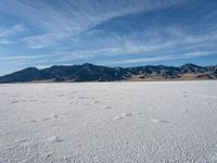 a snow field with mountains in the distance with clouds in the sky above it with footprints across the ice