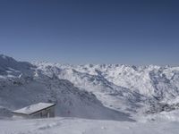 a large mountain range covered in snow and mountains with a sky background, while a person skis down a steep snowy slope with a building below it