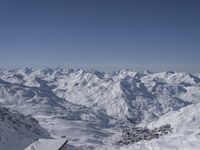 a large mountain range covered in snow and mountains with a sky background, while a person skis down a steep snowy slope with a building below it