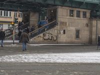 people are walking down the street in the snow by a train station with graffiti on it