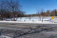 a snowy city street near an empty highway in wintertime with trees and fence, in the foreground