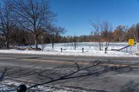 a snowy city street near an empty highway in wintertime with trees and fence, in the foreground