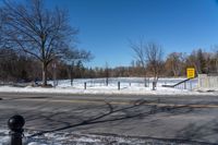 a snowy city street near an empty highway in wintertime with trees and fence, in the foreground