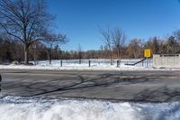 a snowy city street near an empty highway in wintertime with trees and fence, in the foreground