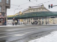 a train passing over a bridge on a snowy day in europe, with people waiting and crossing the road