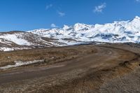 a dirt road winding around a mountain with some snow covered mountains in the background while a small blue sky appears to be on top