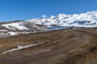 a dirt road winding around a mountain with some snow covered mountains in the background while a small blue sky appears to be on top