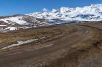 a dirt road winding around a mountain with some snow covered mountains in the background while a small blue sky appears to be on top