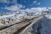 there are snow piled to the ground near mountains and road markings in the foreground