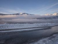 a snowy mountain is reflected on the road by the ocean, and a cloudy blue sky