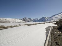 a very long road with some snow on the sides and mountains behind it as seen in this photograph
