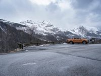 an orange truck is driving down a snowy road with mountains behind it and snow - capped rocks