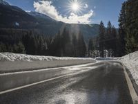 a wet road is surrounded by snow and mountains in the sun, with pine trees on either side