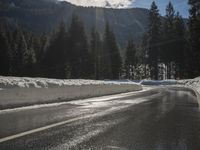 a wet road is surrounded by snow and mountains in the sun, with pine trees on either side