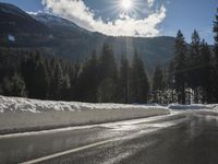 a wet road is surrounded by snow and mountains in the sun, with pine trees on either side