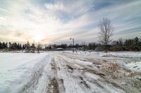 a snow covered country road with bare trees in the background while sunlight is shining through the clouds