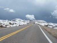 snow covers the roadway and snowy mountains on a sunny day, with a yellow warning sign in front of it