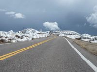 snow covers the roadway and snowy mountains on a sunny day, with a yellow warning sign in front of it