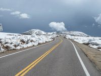 snow covers the roadway and snowy mountains on a sunny day, with a yellow warning sign in front of it