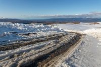 a snowy road lined with snow and rocks leading to mountains in the background, some have tire tracks