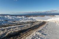 a snowy road lined with snow and rocks leading to mountains in the background, some have tire tracks