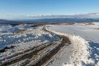 a snowy road lined with snow and rocks leading to mountains in the background, some have tire tracks