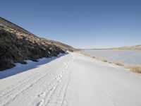 a dirt road next to the snow covered ground with tracks in it and a lone hill in the background