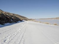 a dirt road next to the snow covered ground with tracks in it and a lone hill in the background