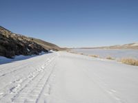 a dirt road next to the snow covered ground with tracks in it and a lone hill in the background